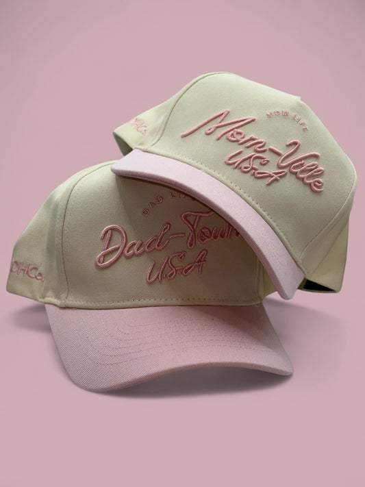 Two Mom-Ville caps arranged together, showing the light pink bills and script fronts