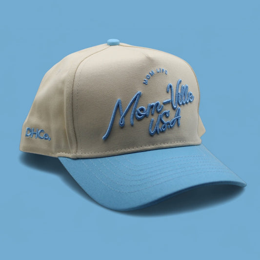 Destination Hat Co. Mom-Ville script cap with beige crown and light blue bill, angled view